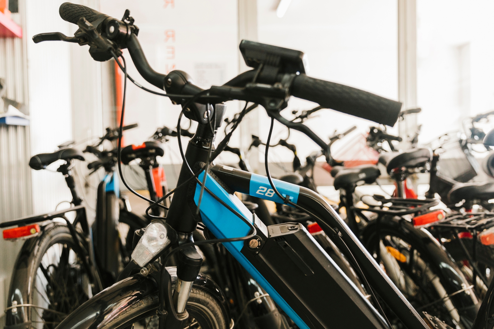 Home 6 Close-up of a blue electric bicycle among other bicycles parked indoors, showing the handlebars, battery, and front light.