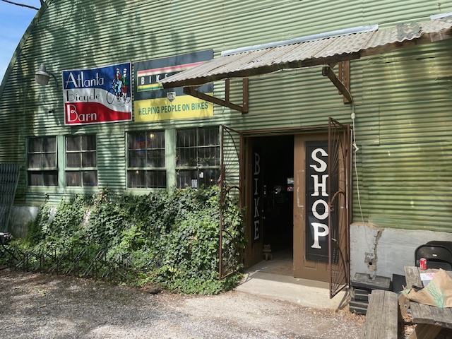 Home 7 Exterior of a green corrugated metal building with signs reading "Atlanta Bicycle Barn" and "Shop," and a yellow banner stating "Helping People on Bikes.