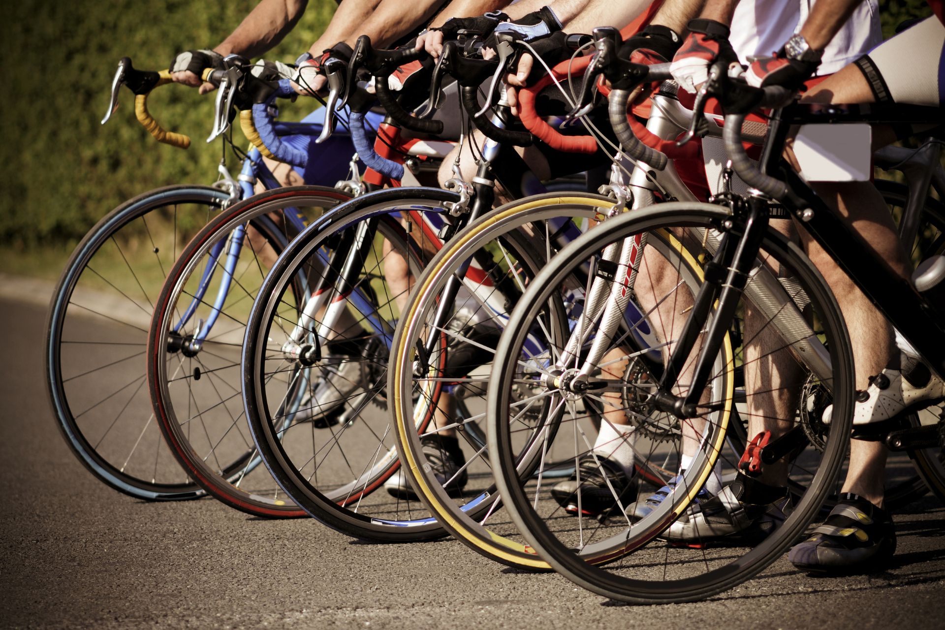Home 3 Close-up of cyclists lined up with their road bikes on an asphalt surface, focusing on the wheels and legs, ready for a race or group ride.