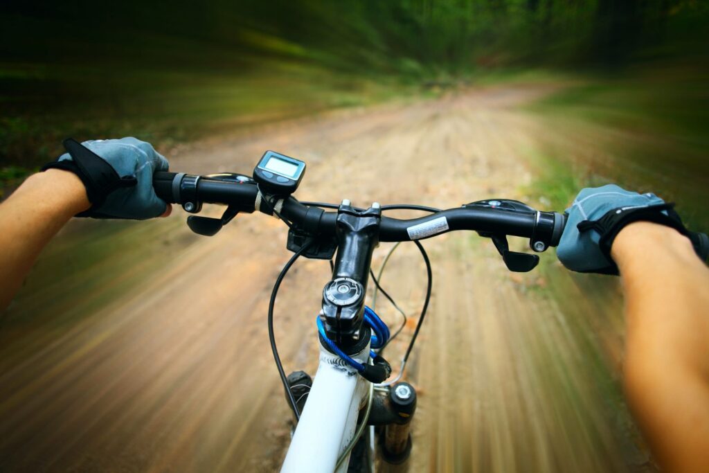 Home 12 A cyclist's hands grip bike handlebars while riding quickly on a dirt path, with a motion blur effect suggesting speed through a forested area.