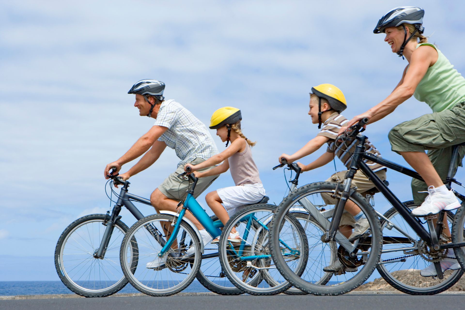Home 5 Four people, two adults and two children, ride bicycles in a line on a paved road. All are wearing helmets, and the sky is partly cloudy.
