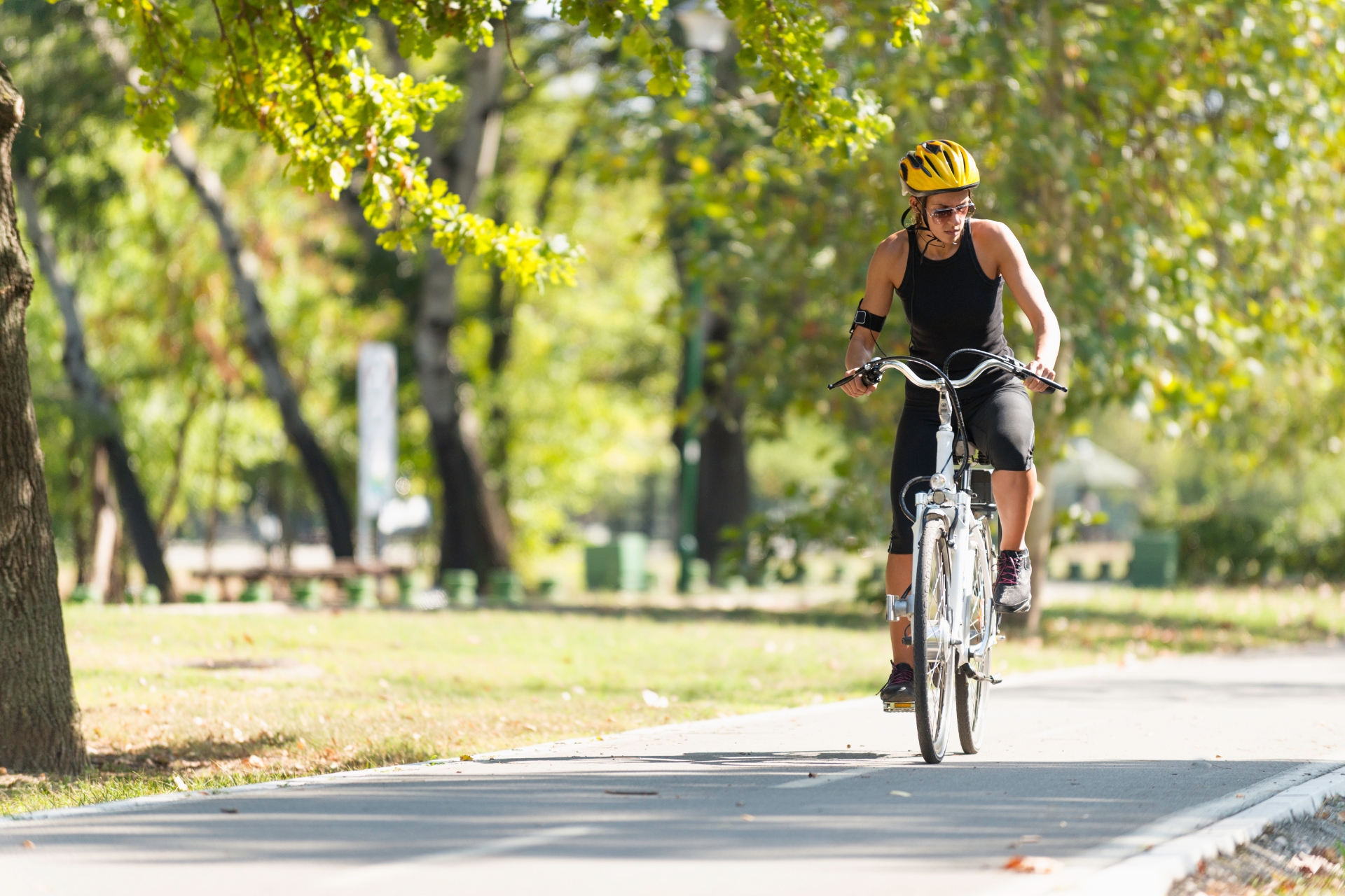 Home 4 A person wearing a yellow helmet and black athletic clothing rides a bicycle on a paved path through a park with green trees.